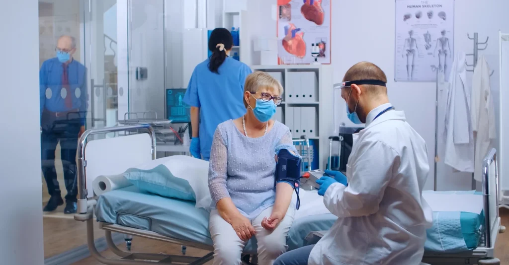 In a bright hospital room, a masked woman sits on her bed as a doctor speaks with her and a nurse works nearby, creating a calm, caring scene.