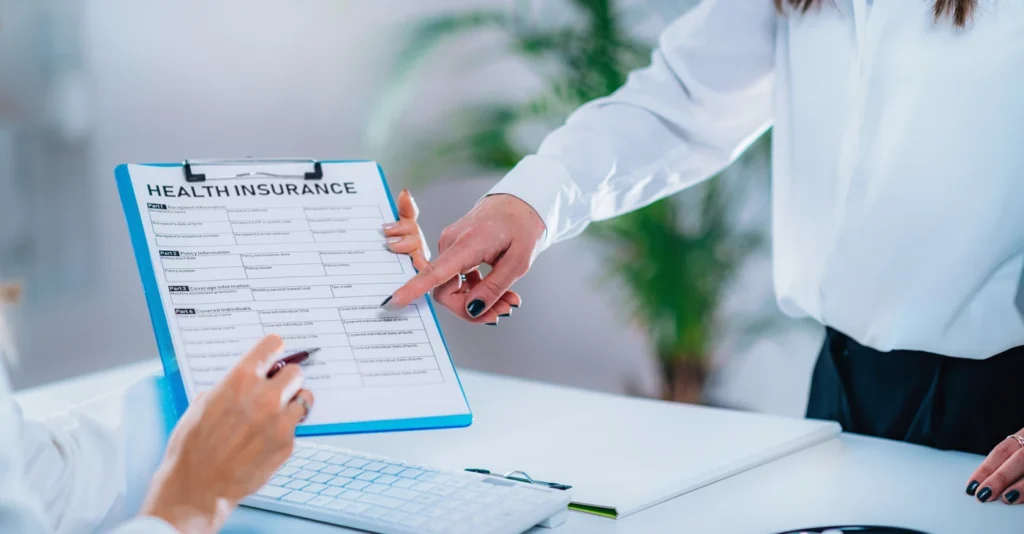 A healthcare worker points to a health insurance form on a blue clipboard as a patient fills it out at a clinic, creating a calm, professional tone.