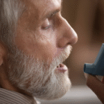 A bearded man with white hair using an asthma inhaler, focusing on his breathing for relief.