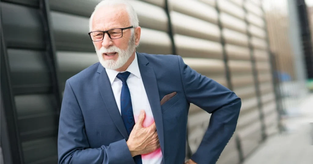 A suited older man clutches his chest, showing signs of chest pain from stress.