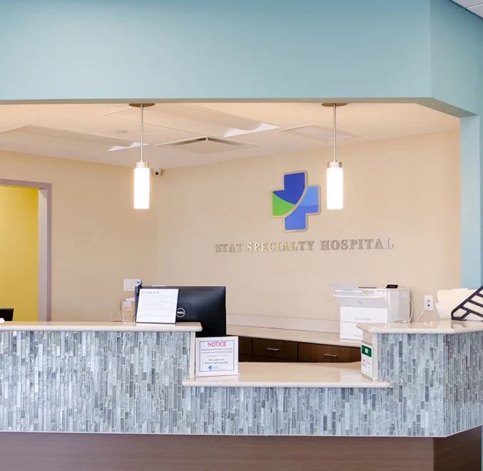 A reception desk at STAT Specialty Hospital. The desk features a modern, textured gray and white tile front with a light-colored countertop. On the back wall, there is a blue and green medical cross logo above the silver lettering "STAT SPECIALTY HOSPITAL." Two cylindrical pendant lights hang from the ceiling, and a computer monitor and printer are visible behind the counter. The walls are a mix of light tan and soft blue.