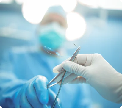 A close-up, focused shot of a surgical hand-off in an operating room. A gloved hand in white passes a metal surgical instrument to another gloved hand in blue. The background shows a surgeon in a mask and cap, softly blurred under bright surgical lights.