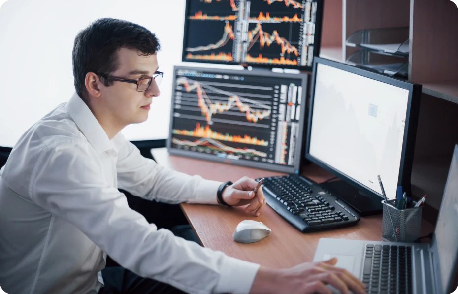 A man in glasses is seated at a desk, using two computers.