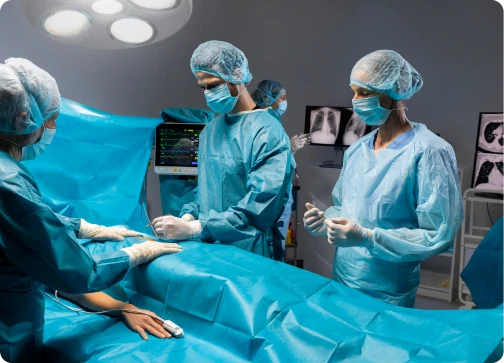 A team of surgeons and medical staff in blue scrubs, masks, and hairnets performing a procedure in an operating room. They are gathered around a patient covered in a blue surgical drape under bright overhead medical lights, with heart monitors and X-ray scans visible in the background.