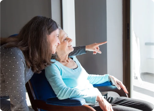 A woman gestures towards an elderly woman sitting comfortably in a chair.