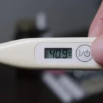 A close-up of a digital thermometer displaying a fever-grade temperature, held by a person in a soft-lit, comfortable bedroom setting.