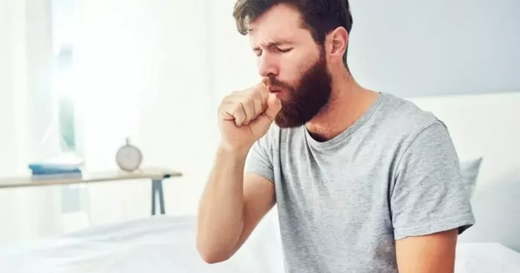 A photograph of a bearded man in a grey t-shirt sitting on a bed and coughing into his fist. He has his eyes closed in discomfort, set against a bright, blurred bedroom background.