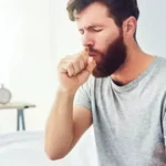 A photograph of a bearded man in a grey t-shirt sitting on a bed and coughing into his fist. He has his eyes closed in discomfort, set against a bright, blurred bedroom background.