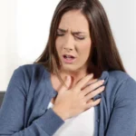 Woman sitting on a grey sofa indoors, holding her chest with a pained expression, illustrating chest tightness or shortness of breath.