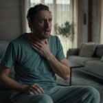 A man sitting in a living room with a distressed expression, holding his hand to his throat.