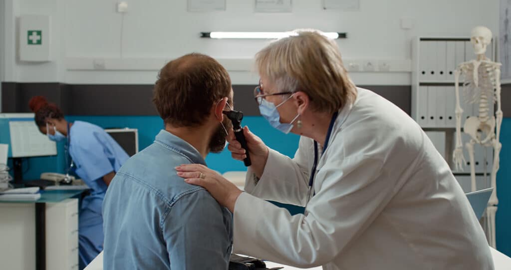 Doctor examining a patient’s ear with an otoscope in a medical clinic while another healthcare worker is visible in the background.