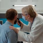 Doctor examining a patient’s ear with an otoscope in a medical clinic while another healthcare worker is visible in the background.