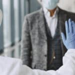 A medical professional in a hazmat suit and blue gloves holding their hand up to signal a man in a face mask to stop.