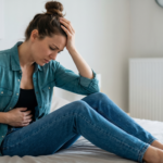 A woman sitting on a bed, wearing blue jeans and a teal shirt, holding her head and stomach as if experiencing pain or discomfort.