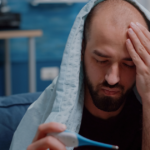 Man with a fever checking his temperature under a blanket.