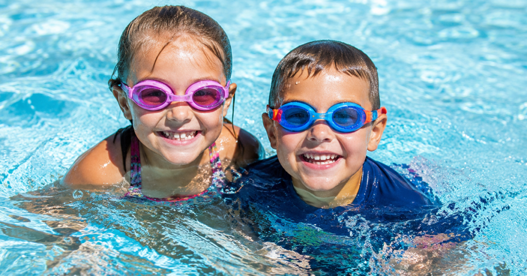Two children wearing goggles smiling in a swimming pool.