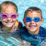 Two children wearing goggles smiling in a swimming pool.