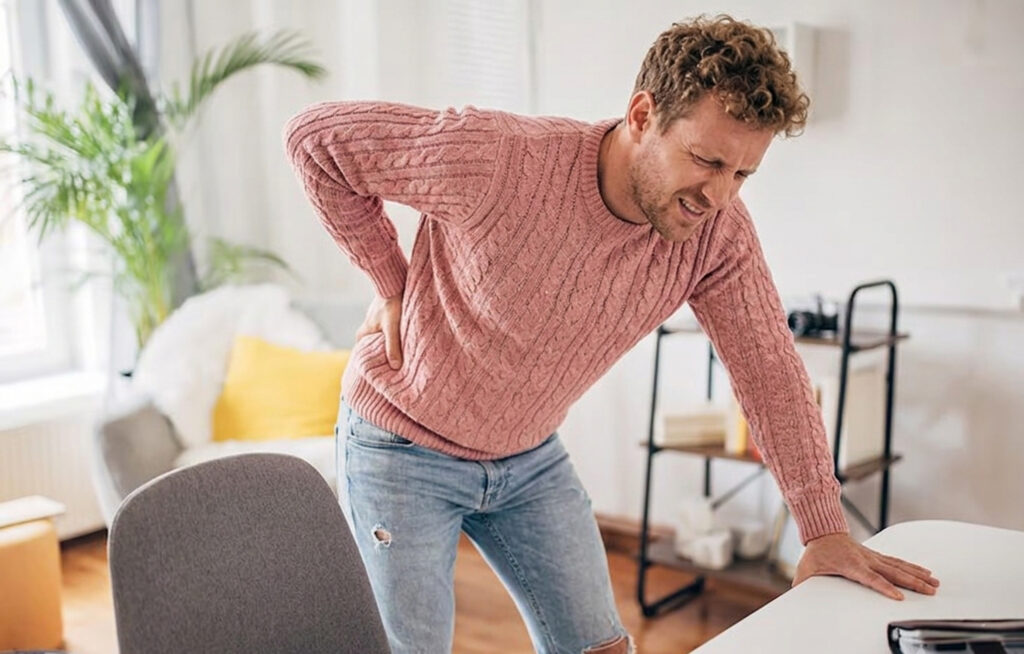 A man in a pink knit sweater leaning over a table while holding his lower back in pain.