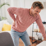 A man in a pink knit sweater leaning over a table while holding his lower back in pain.