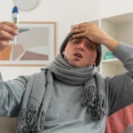 Sick man sitting on a couch checking a digital thermometer for a fever.