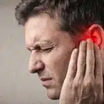 A man grimacing in pain while holding his ear, which is highlighted in red to indicate discomfort or injury.