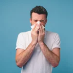 A man blowing his nose into a tissue against a blue background.