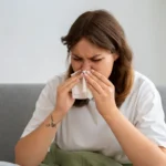 Woman sitting on a sofa blowing her nose with a tissue.
