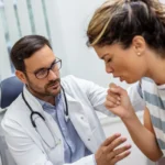 Male doctor wearing glasses examines a coughing female patient indoors.