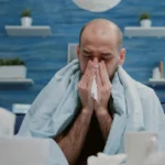 Sick man wrapped in blanket blowing nose with tissue paper.