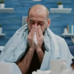 Sick man wrapped in blanket blows nose with paper tissue.