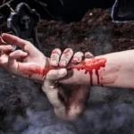 Pale zombie hand grabbing bloody arm above misty grave site.
